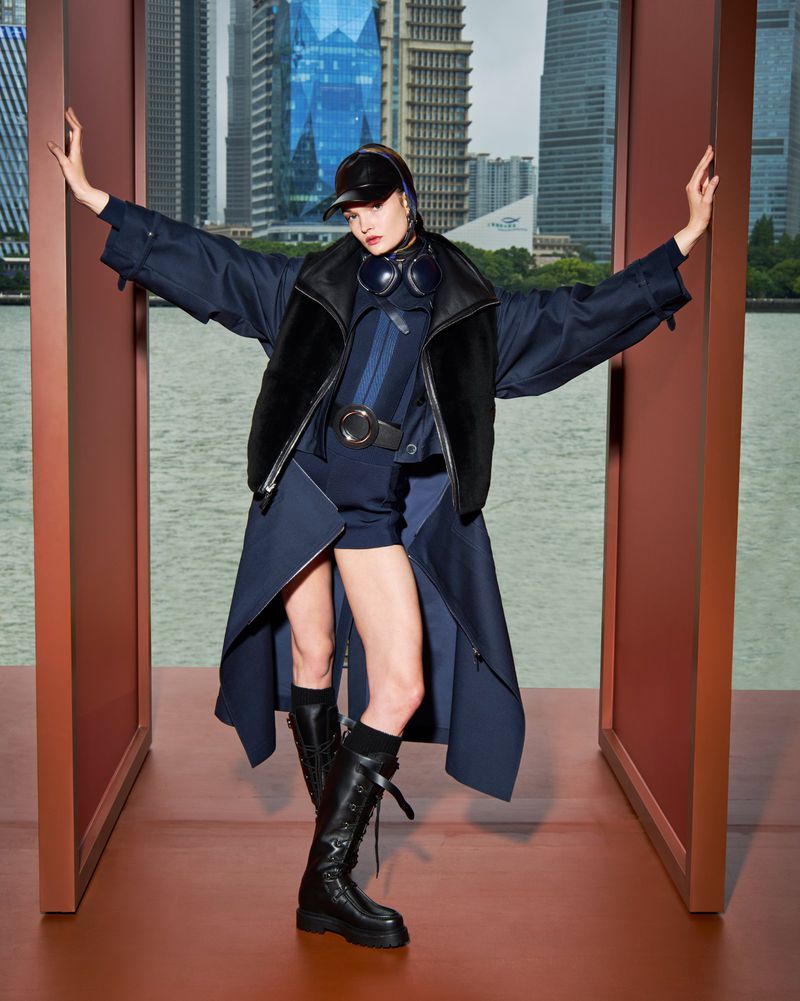 Fashionable woman in navy outfit and boots strikes a pose with city skyline in background, framed by architectural elements on a studio set.