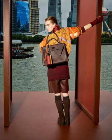 Fashion shot of a woman posing outdoors in leather shorts and boots, holding a handbag, with an urban skyline and harbor in the background.