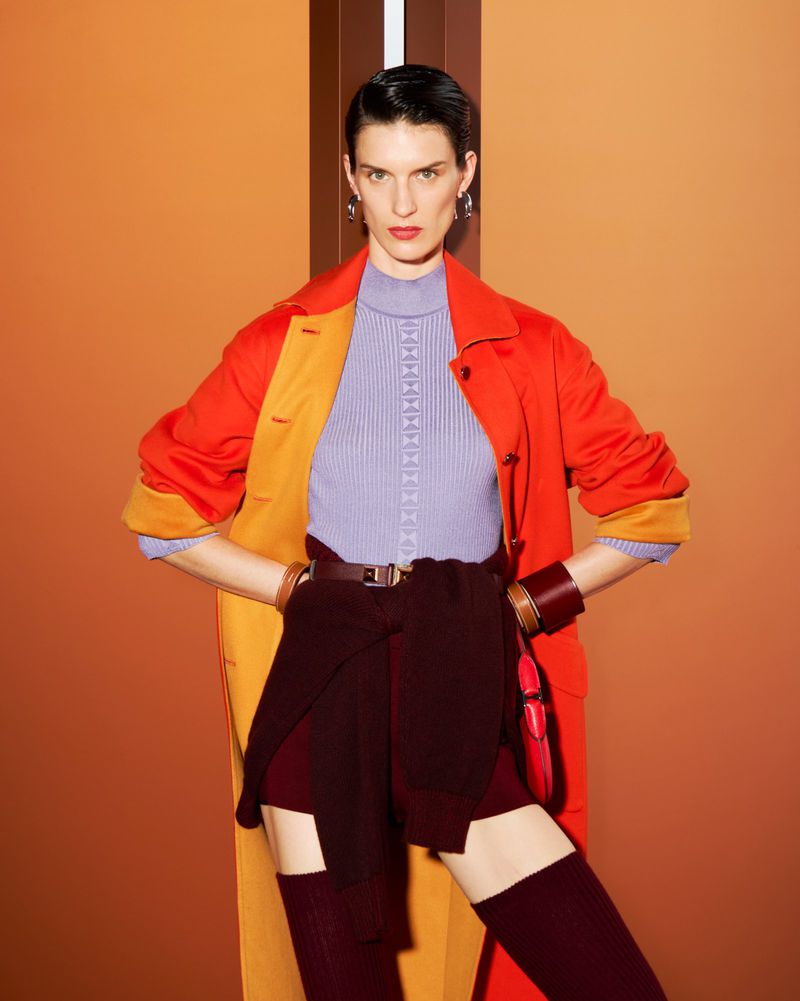 A woman poses in an orange coat, purple knit top, burgundy shorts, and knee-high socks against an orange background in a fashion shot.