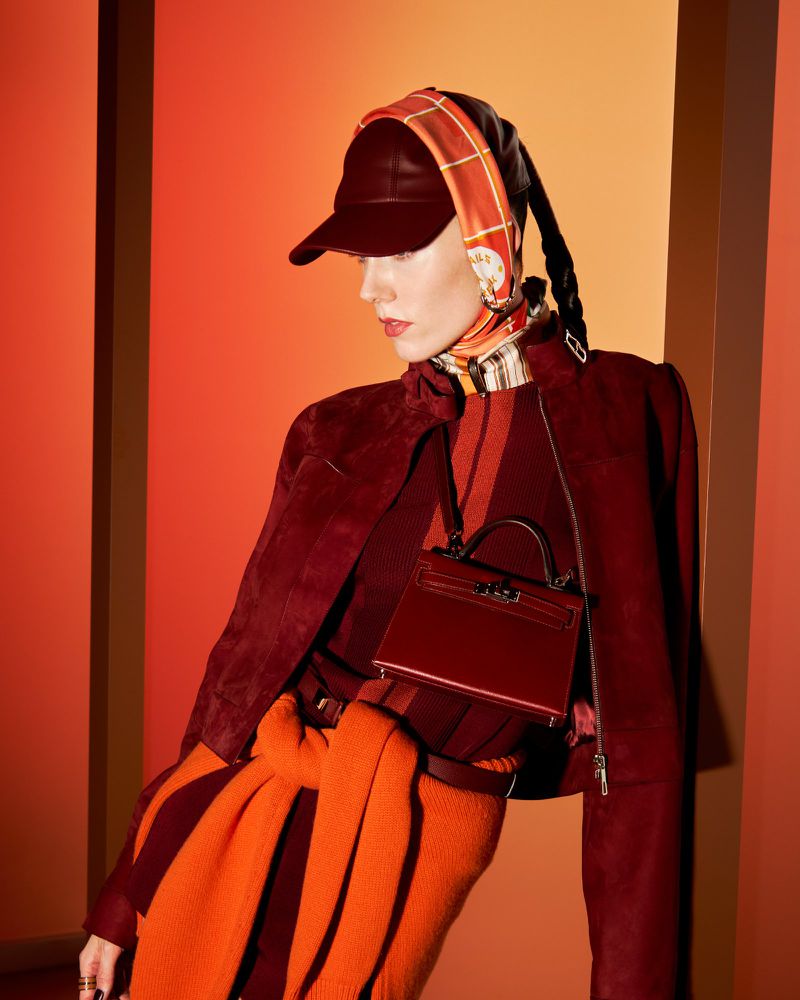 Fashionable adult female poses in a studio wearing a burgundy jacket, striped top and skirt, matching headscarf and cap, and small purse.