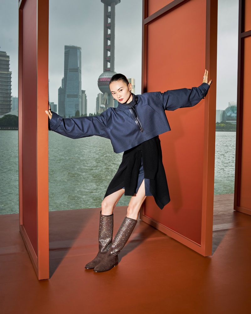 Fashion shot of a model in a blue top, black skirt, and glitter boots, posing in front of the Shanghai skyline on a cloudy day.
