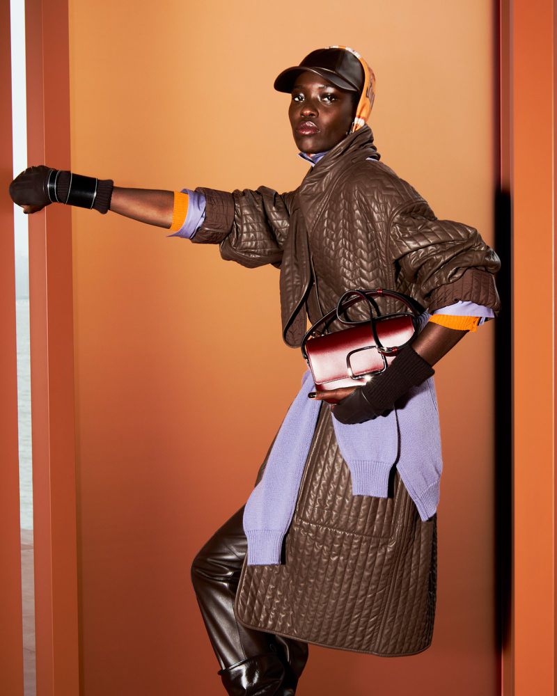 A Black woman models fashion with a brown quilted jacket, black cap, gloves, brown leather pants, and purse against an orange background.