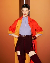 Model Marte Mei van Haaster in an orange coat, lilac top, and burgundy tied knitwear with hands on hips against a warm background.