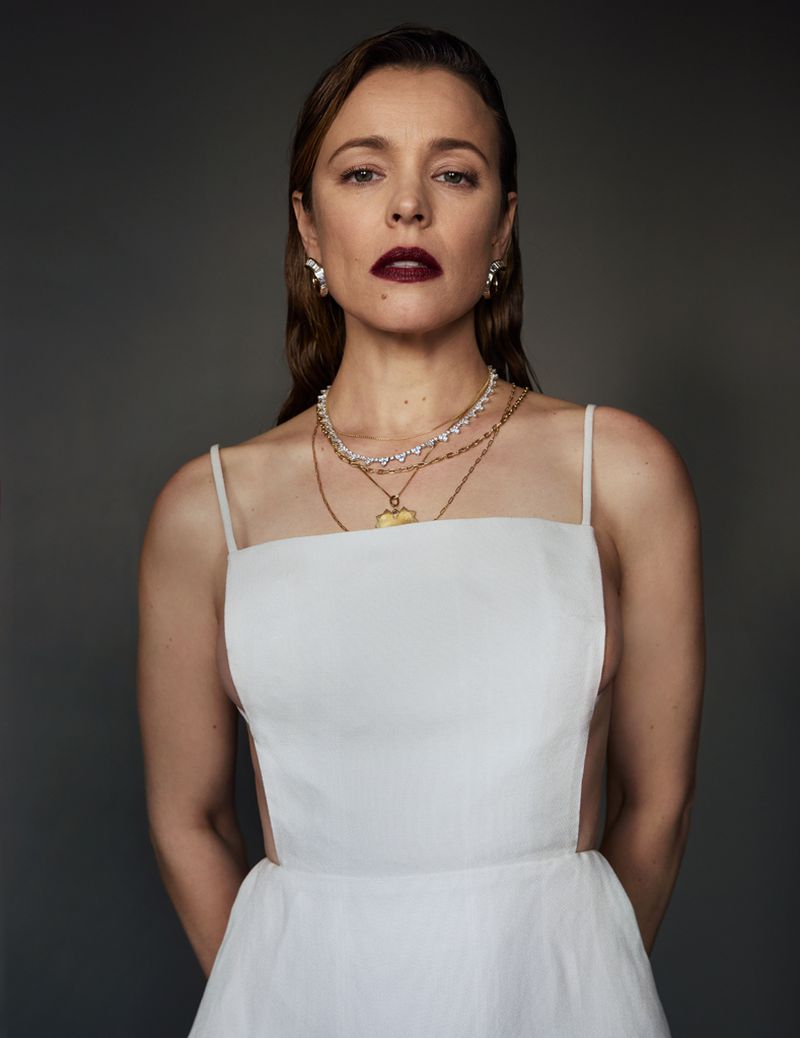 Rachel McAdams in a white open-back dress, layered gold necklaces, and bold burgundy lipstick against a seamless dark grey studio backdrop.