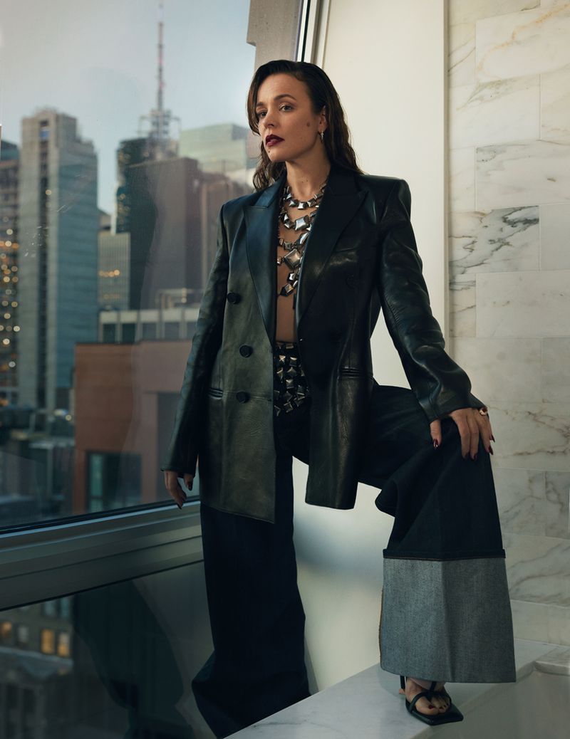 Rachel McAdams poses in an oversized black leather blazer and wide-leg trousers beside a high-rise window overlooking city buildings.