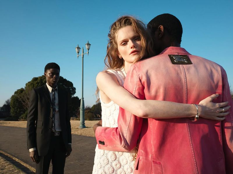 Lily Chapman in a textured white dress embraces a man in a pink jacket outdoors while a man in a black suit stands in the background.