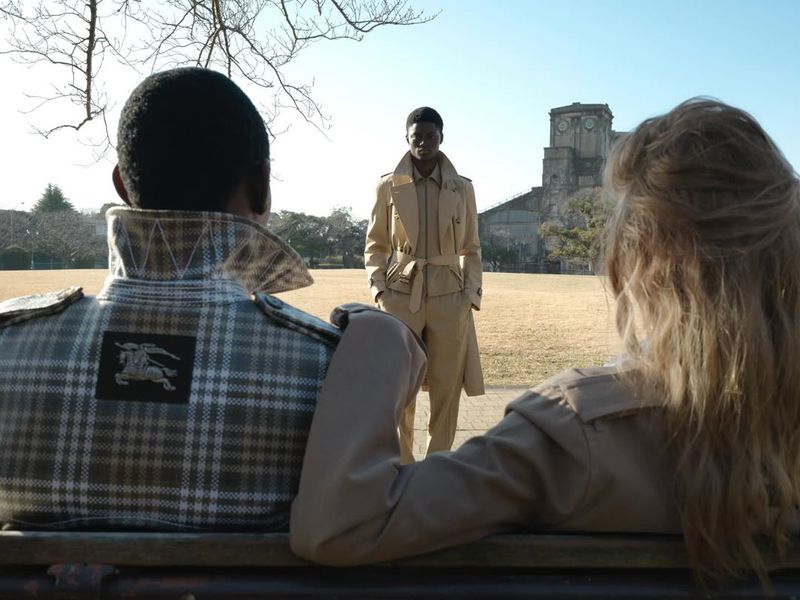 Model in a belted beige trench coat standing in an open field framed by two seated figures wearing coats on a park bench.