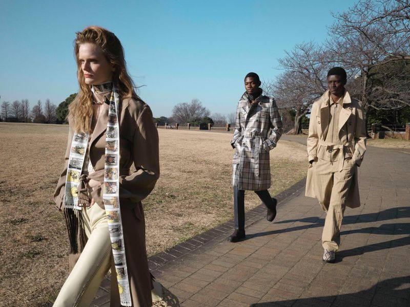 Three fashion models in trench coats walking along a paved park pathway under a clear blue sky.