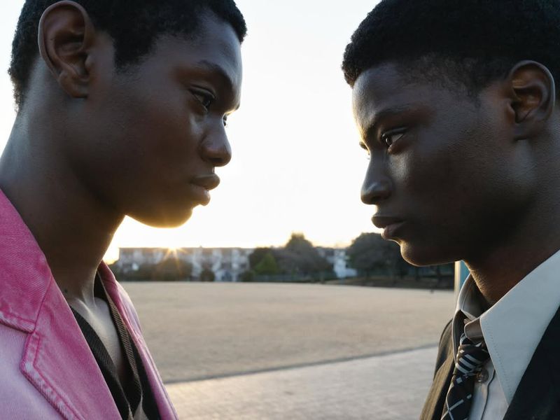 Close profile shot of two Black men facing each other outdoors in front of an open field during sunset.