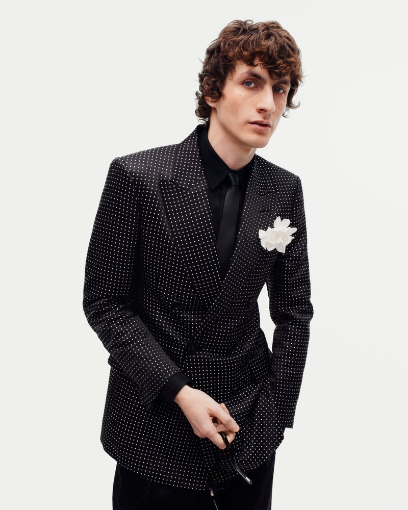 Male model in a black double-breasted polka dot blazer with a black tie and white flower pin against a white backdrop.