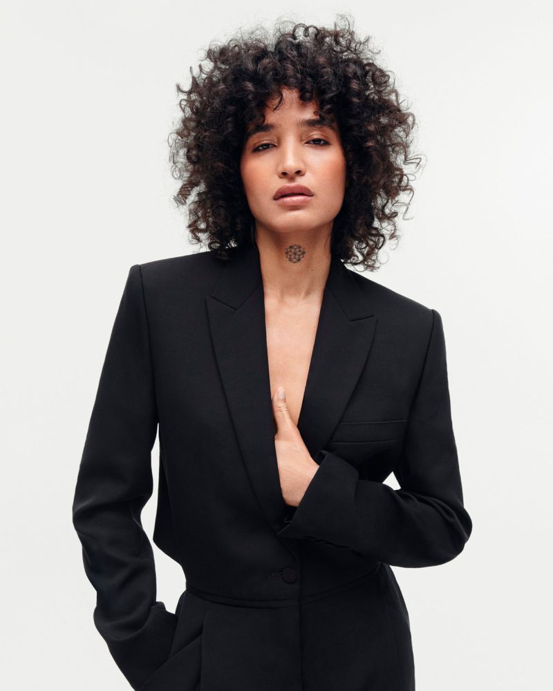 Indya Moore wearing a tailored black cropped blazer against a white studio background.