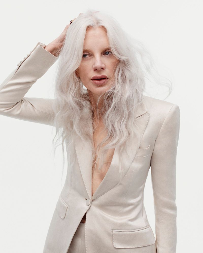 Kristen McMenamy with long white hair posing in a tailored cream satin blazer against a white seamless background.