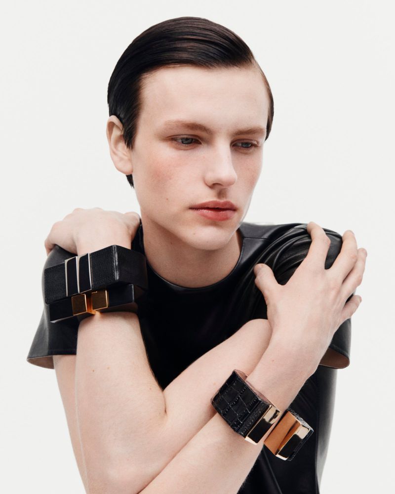 Model Henry Kitcher wearing a black leather top and chunky cuffs with crossed arms against a white seamless background.
