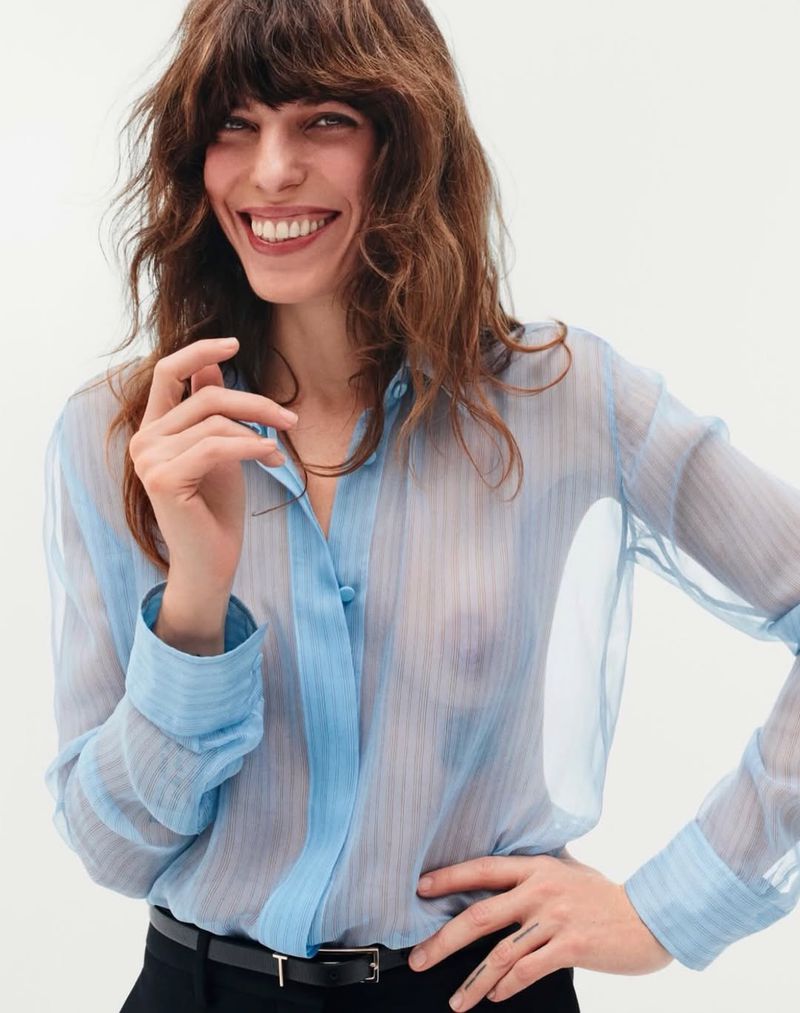 Lou Doillon smiling in a sheer light blue button-down shirt and black belted trousers against a white studio background.