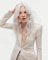 Kristen McMenamy with long white hair posing in a tailored cream satin blazer against a white seamless background.