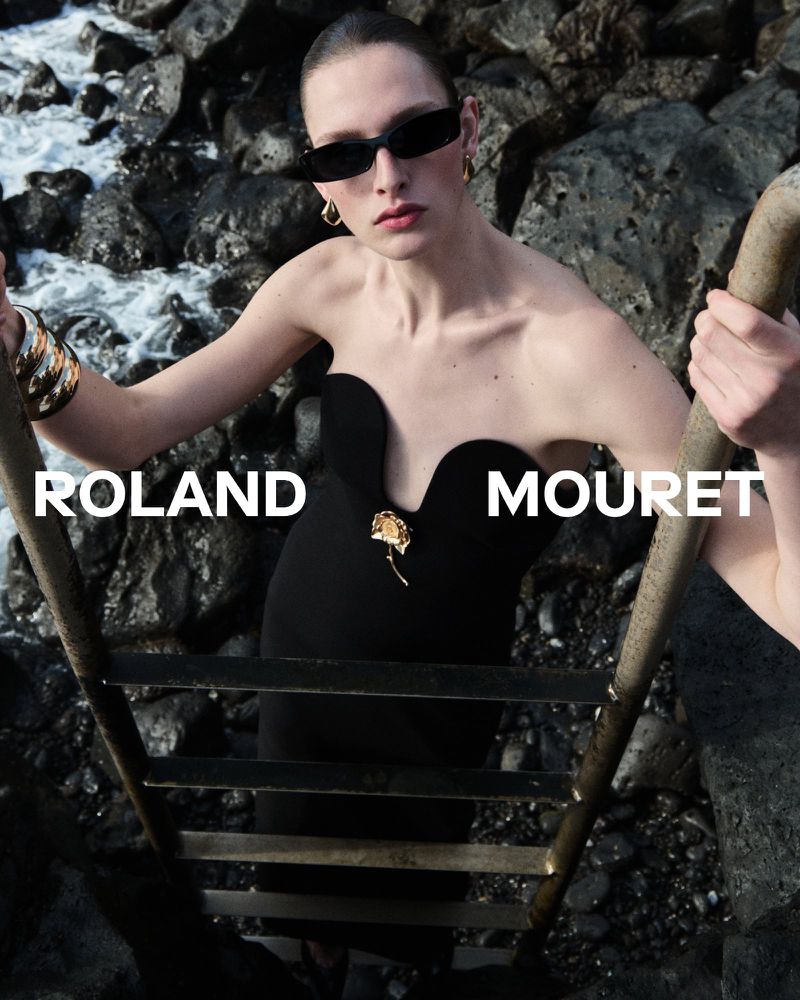 Model Anna Rud wearing a black strapless Roland Mouret dress and sunglasses while holding a ladder against a rocky coastal backdrop.