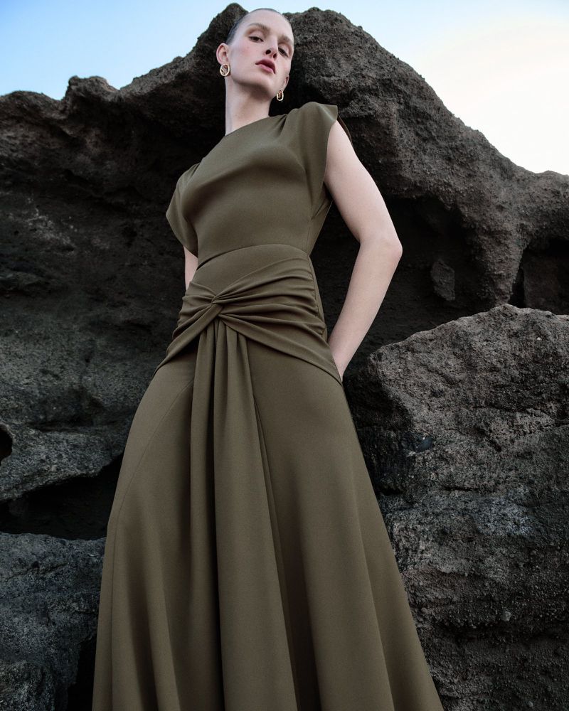 Model Anna Rud wearing an olive green draped evening gown posing against dark volcanic rocks.