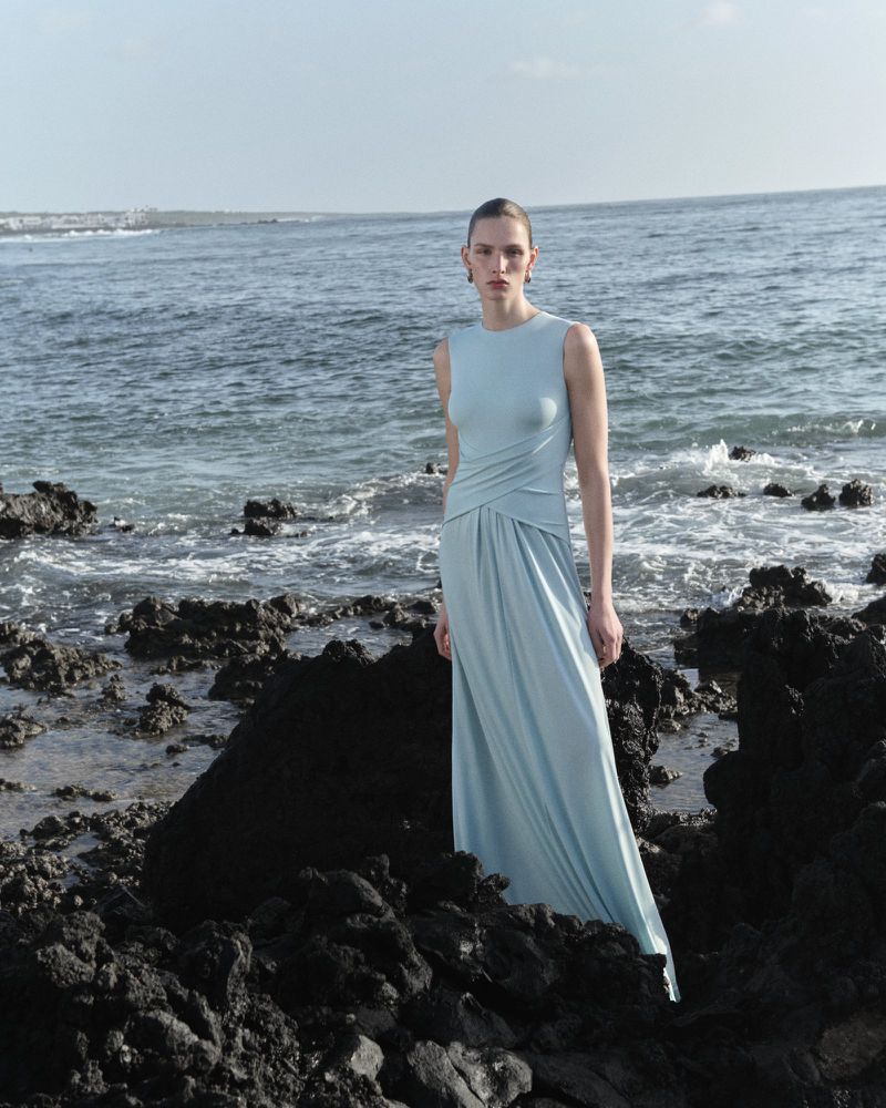 Model Anna Rud wearing a sleeveless light blue draped gown standing on dark coastal rocks by the ocean.