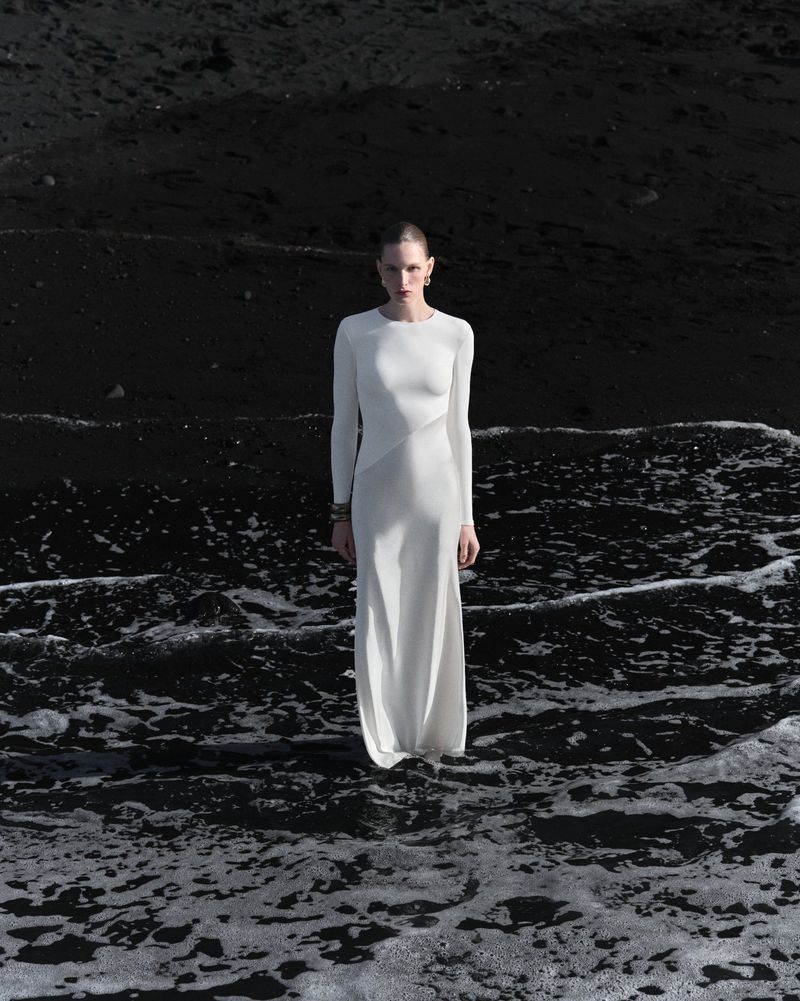 Model in a long white evening dress standing on a dark volcanic beach with ocean waves for Roland Mouret