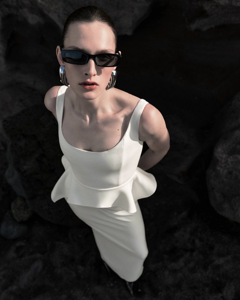 High-angle view of a woman wearing a white peplum dress, dark sunglasses, and silver hoop earrings against dark rocks.