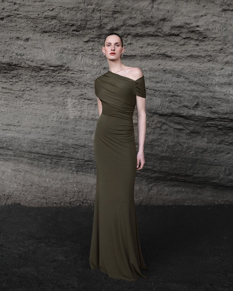 Model Anna Rud poses in an olive green one-shoulder draped evening gown against a dark textured rock wall.