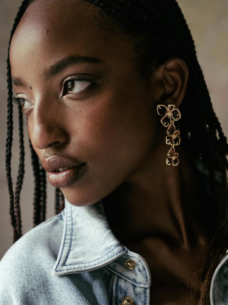 Model Divine Mugisha wearing gold floral drop earrings and a light wash denim jacket with braided hair.