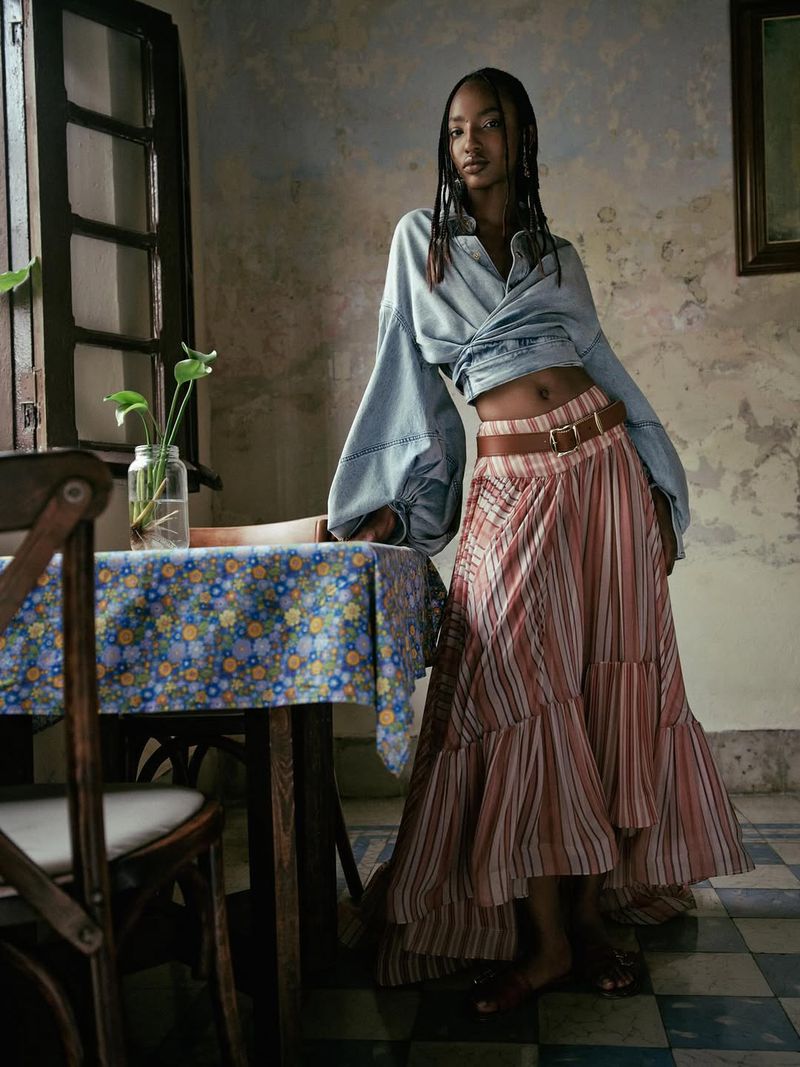 Model Divine Mugisha posed in a cropped denim wrap top and tiered striped maxi skirt beside a floral-draped table.