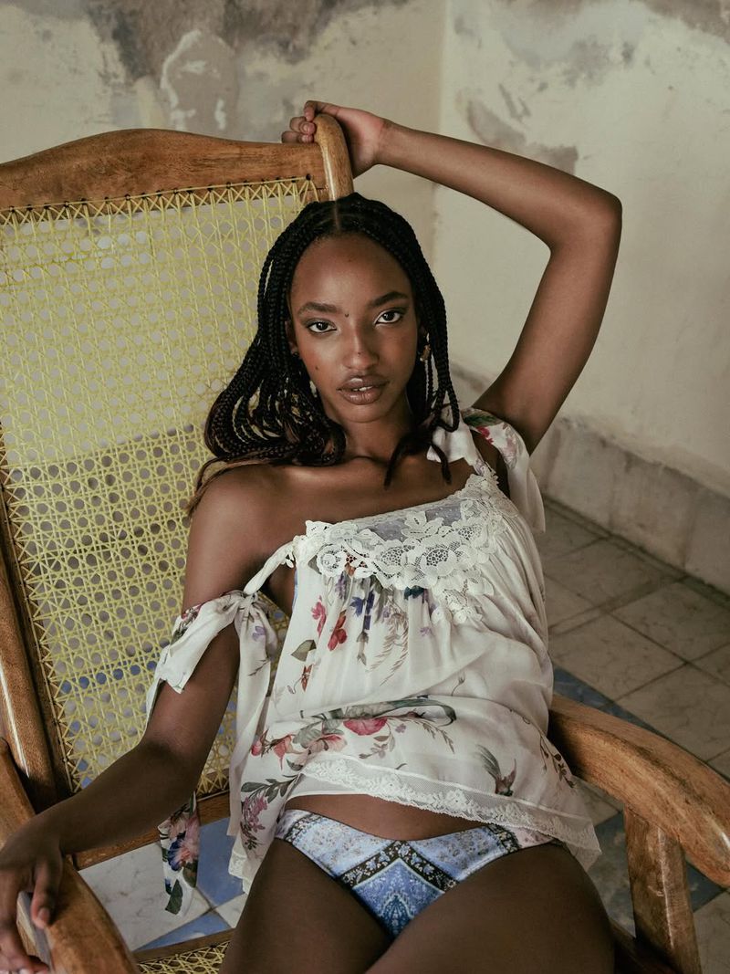 Divine Mugisha reclined in a wooden woven chair wearing an off-the-shoulder floral lace top and patterned briefs.