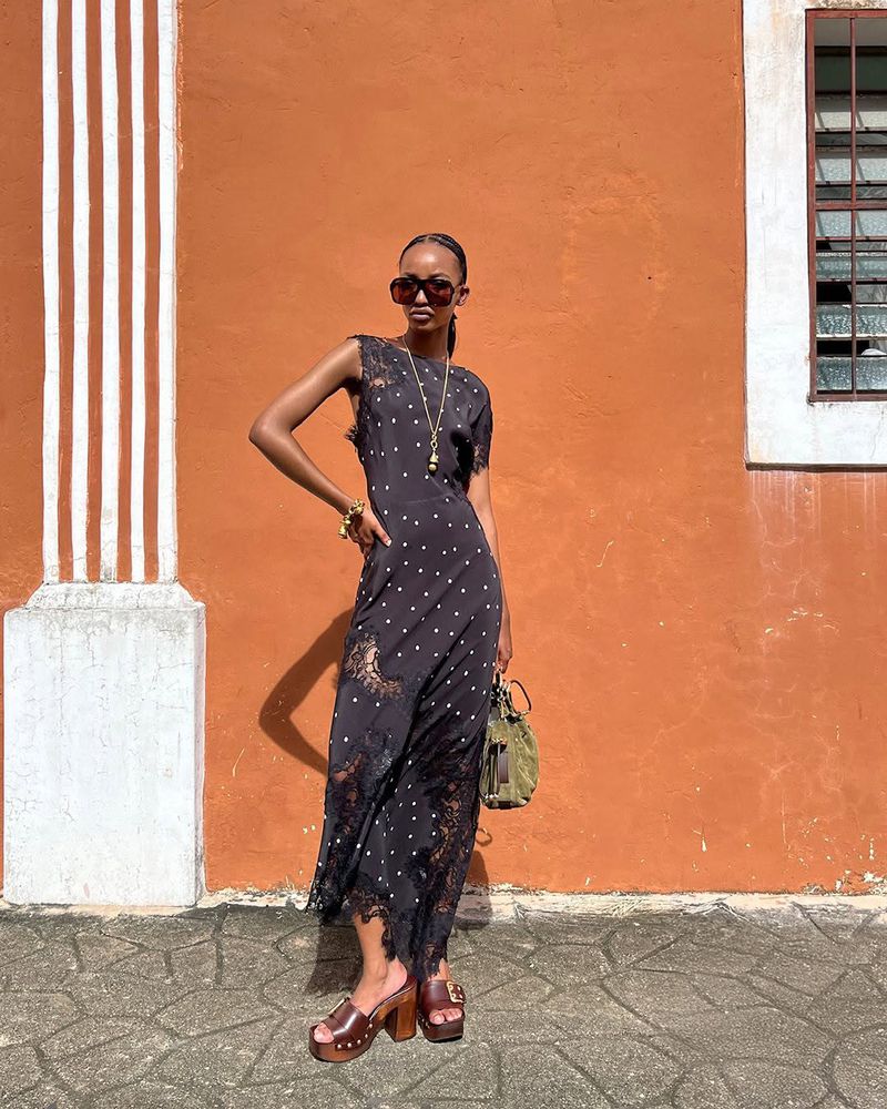 Model Divine Mugisha in a black polka dot lace maxi dress and platform clogs standing against a terracotta wall in bright sunlight.