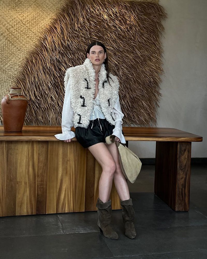 Giedre Dukauskaite in a white shearling vest and black shorts seated on a wooden table for Shopbop Spring 2026.