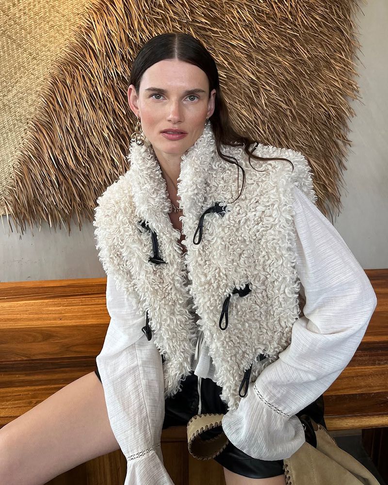 Model Giedre Dukauskaite wearing a textured cream fleece vest and white blouse, seated in front of a woven straw backdrop.