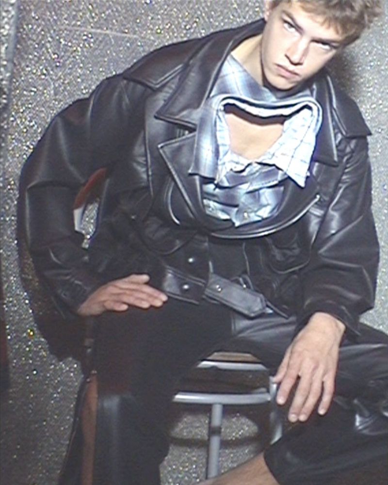 Male model seated wearing an oversized black leather jacket and cut-out collar shirt against a glittery textured silver wall.