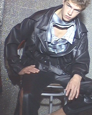 Male model seated wearing an oversized black leather jacket and cut-out collar shirt against a glittery textured silver wall.