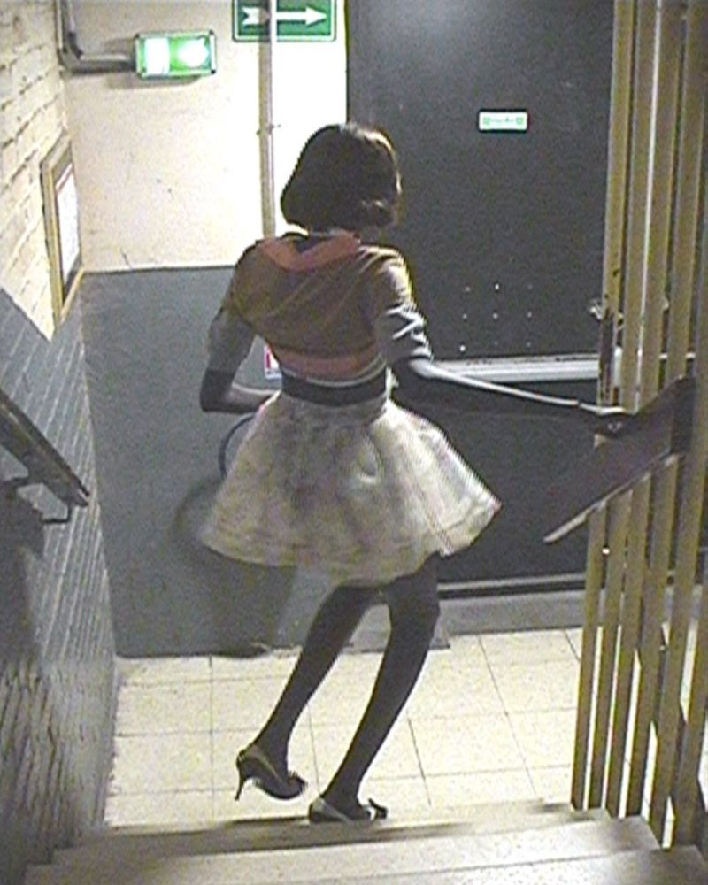 Model in a white tulle skirt, cropped jacket, and black tights standing on a stairwell landing viewed from a high angle.