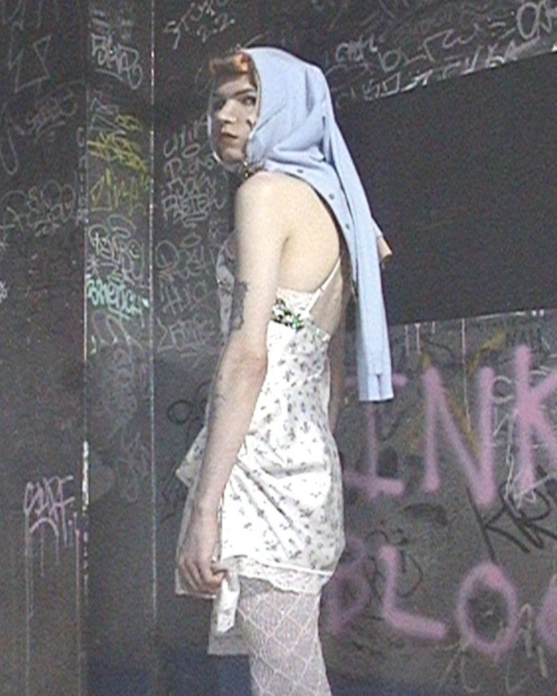 Model wearing a light blue hood, white floral slip dress, and lace tights standing against a graffiti-covered wall.