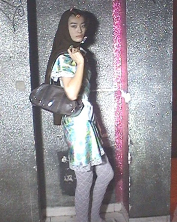 Model in a patterned silk mini dress, textured grey tights, and dark headscarf holding a brown handbag against a textured wall.