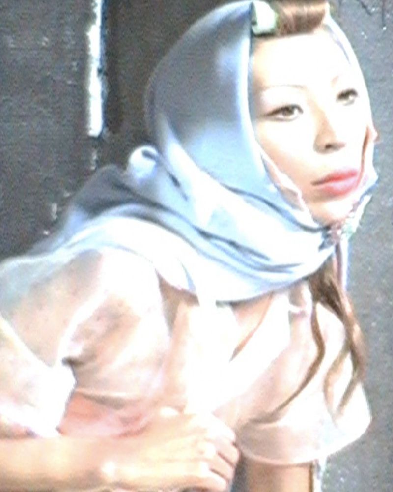An East Asian woman wearing a pale blue headscarf and rolled bangs looking aside against a dark wall.