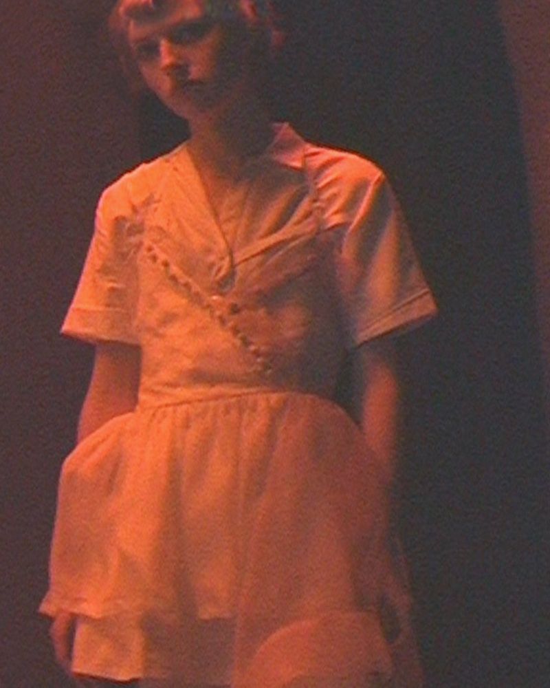 Model wearing a short-sleeved collared dress under warm ambient lighting against a dark background.