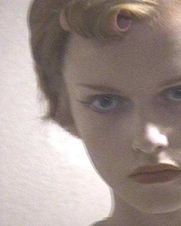 Close-up portrait of a woman with styled vintage curls, winged eyeliner, and a pink hair roller visible in her hair.