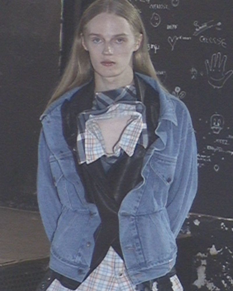 Model with long blonde hair wearing a blue denim jacket over a layered plaid shirt and black vest against a chalkboard wall.