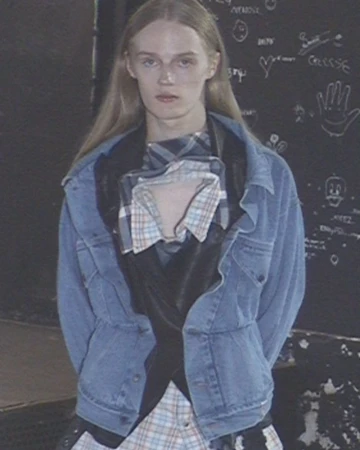 Model with long blonde hair wearing a blue denim jacket over a layered plaid shirt and black vest against a chalkboard wall.
