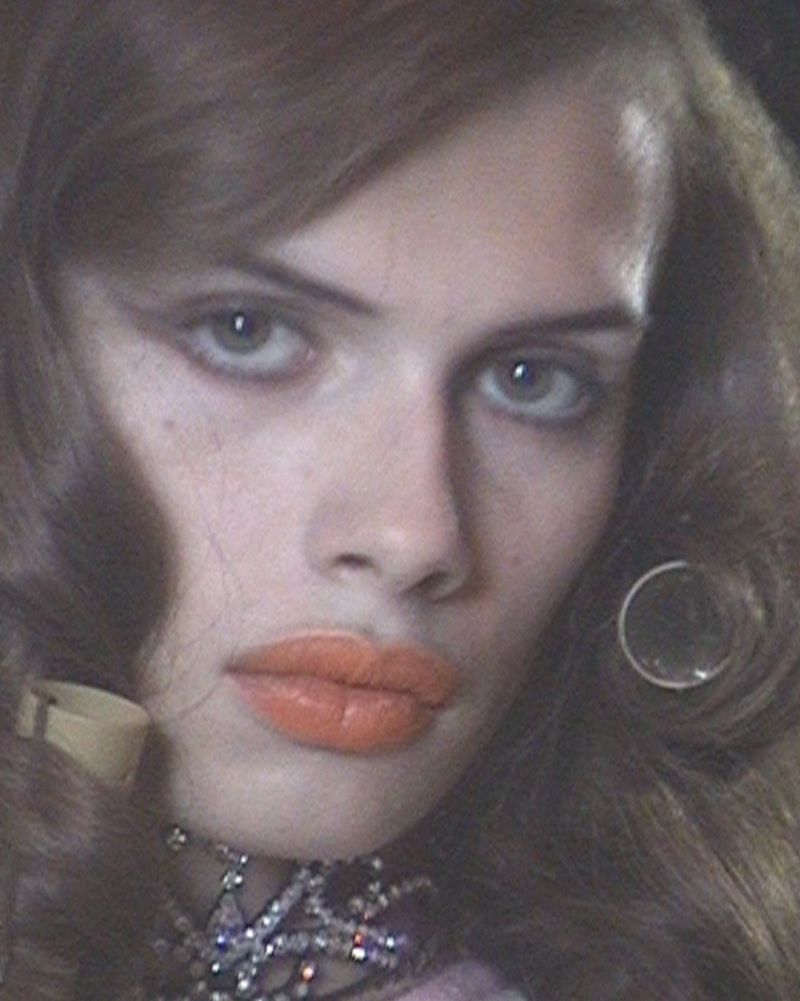 Close-up portrait of a model with voluminous brunette hair, peach lipstick, a round earring, and a crystal choker necklace.