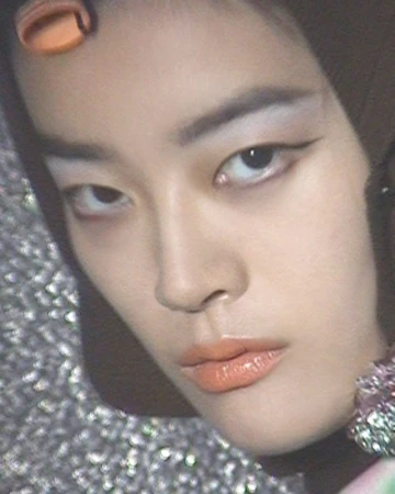 Close-up portrait of an East Asian model wearing an orange hair roller, dark headwrap, and winged eyeliner against a glittery backdrop.