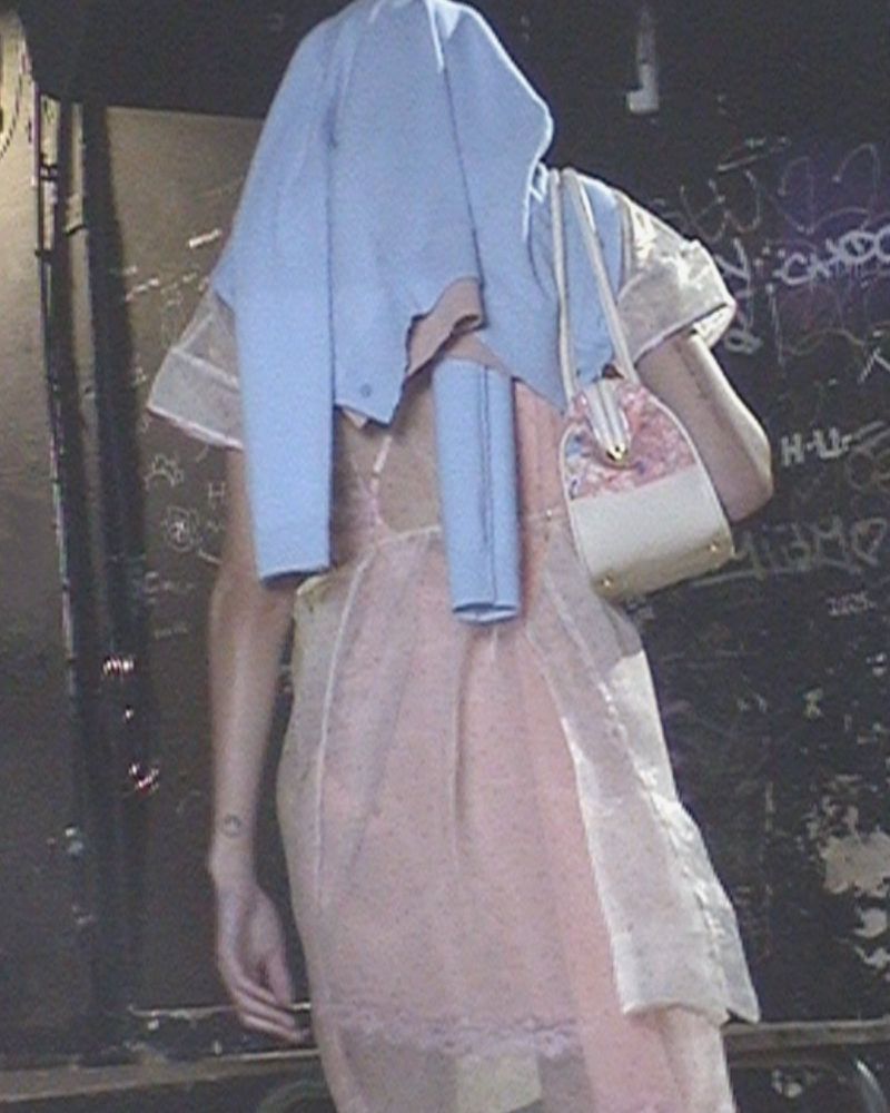 Back view of a person with a light blue sweater draped over their head, wearing a sheer pink slip dress and carrying a shoulder bag.