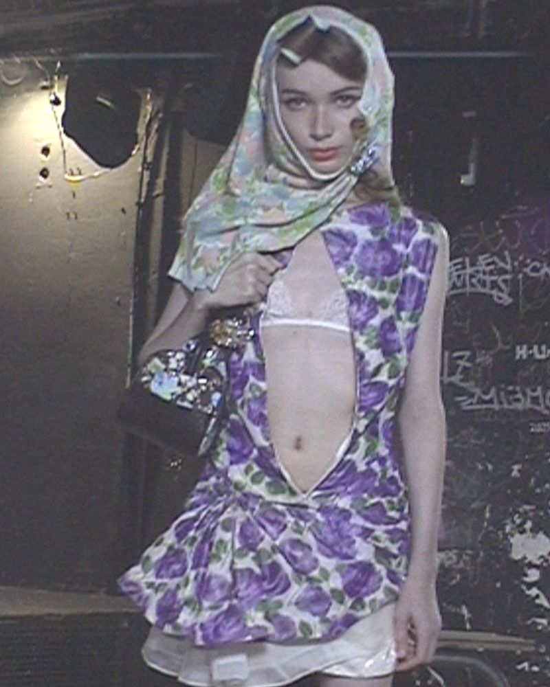 Model wearing a floral babushka headscarf and unzipped purple floral mini dress against a dark graffiti wall.
