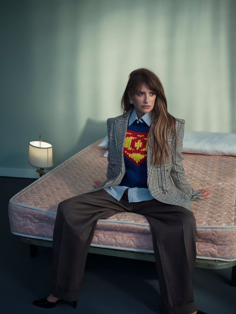 Penélope Cruz sits on a bare mattress wearing a plaid blazer, patterned knit sweater, and wide-leg trousers.