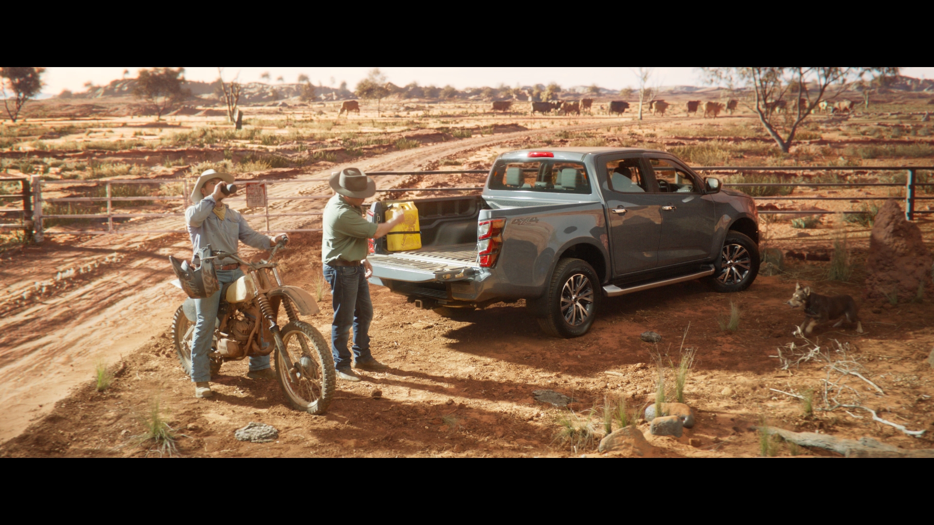 Two men by a gray pickup truck parked on a dirt road in a rural landscape, with cattle grazing in the background on a sunny day.