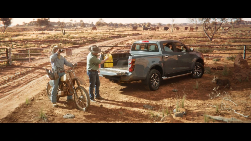 Two men by a gray pickup truck parked on a dirt road in a rural landscape, with cattle grazing in the background on a sunny day.