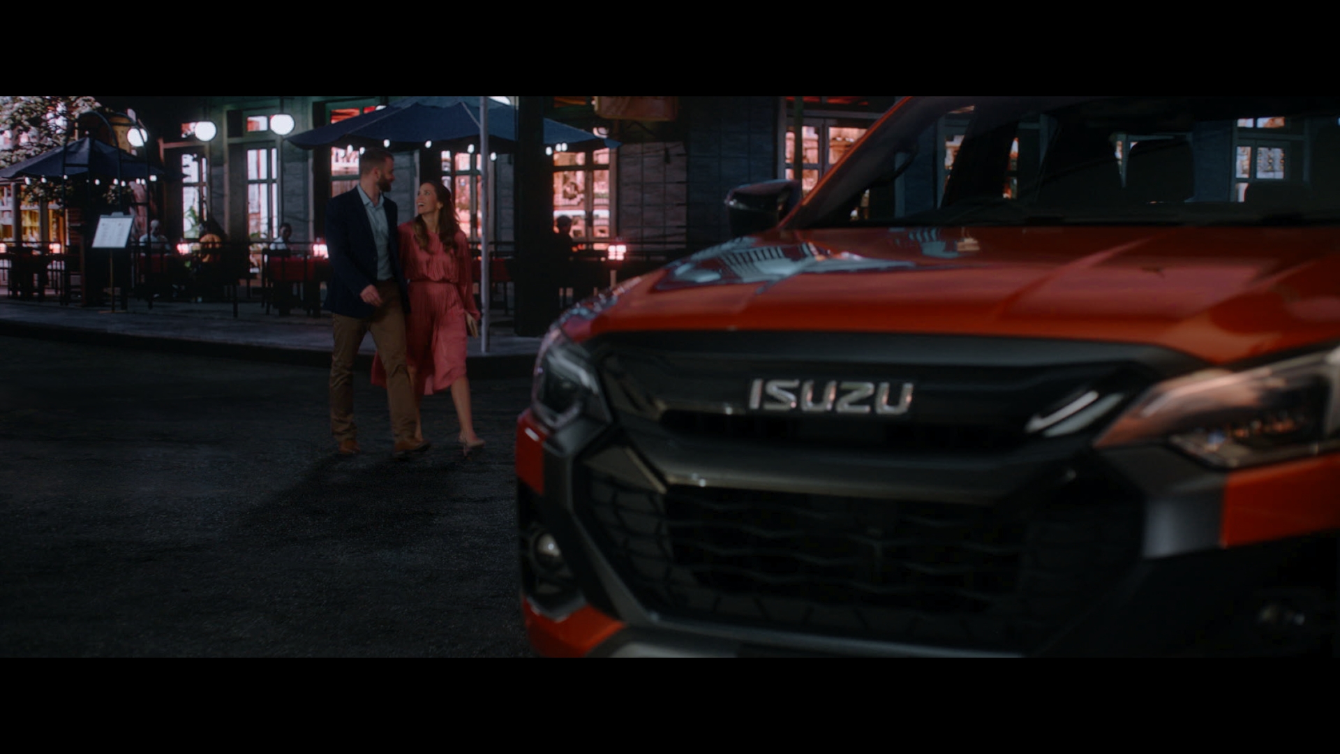 Isuzu truck parked on a city street at night, with a couple walking in the background near outdoor cafe seating.