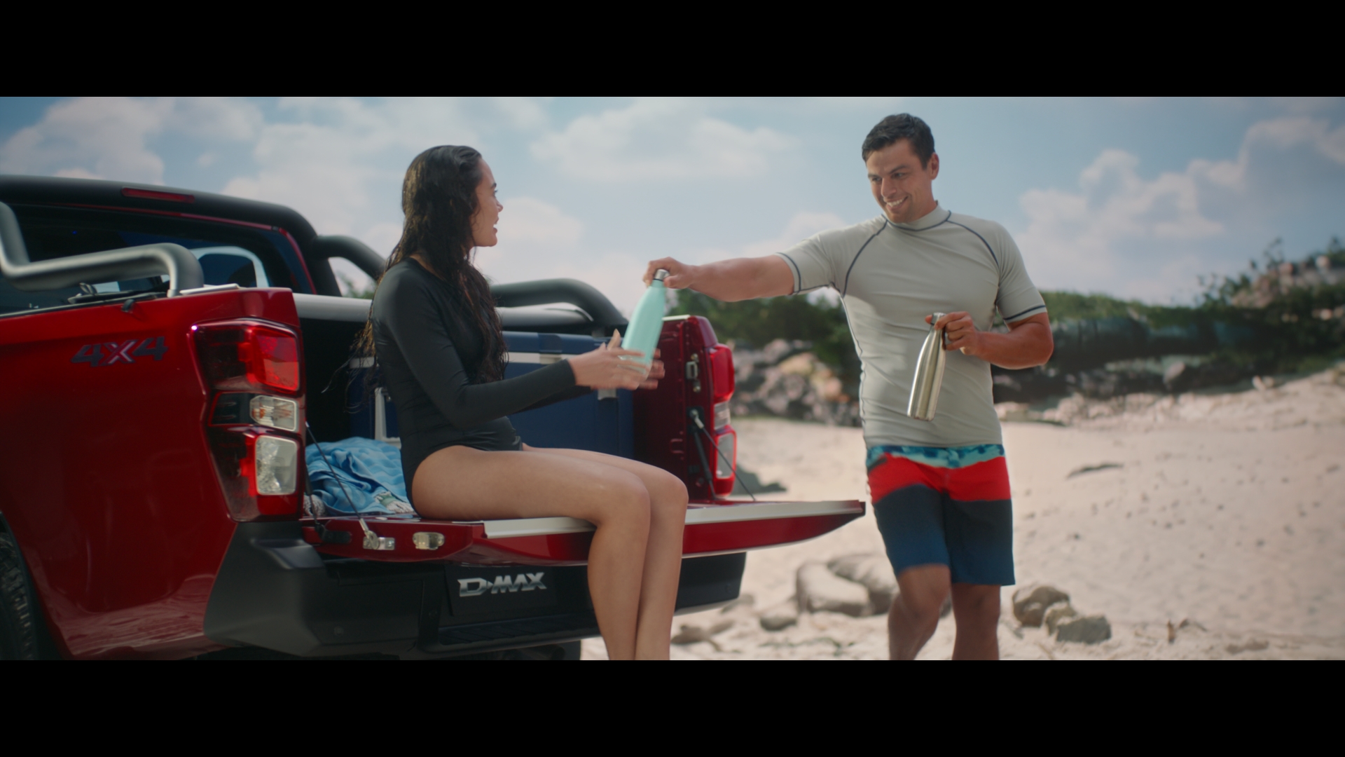 Man hands a water bottle to a woman sitting on the tailgate of a red pickup truck on a sandy beach under a blue sky.